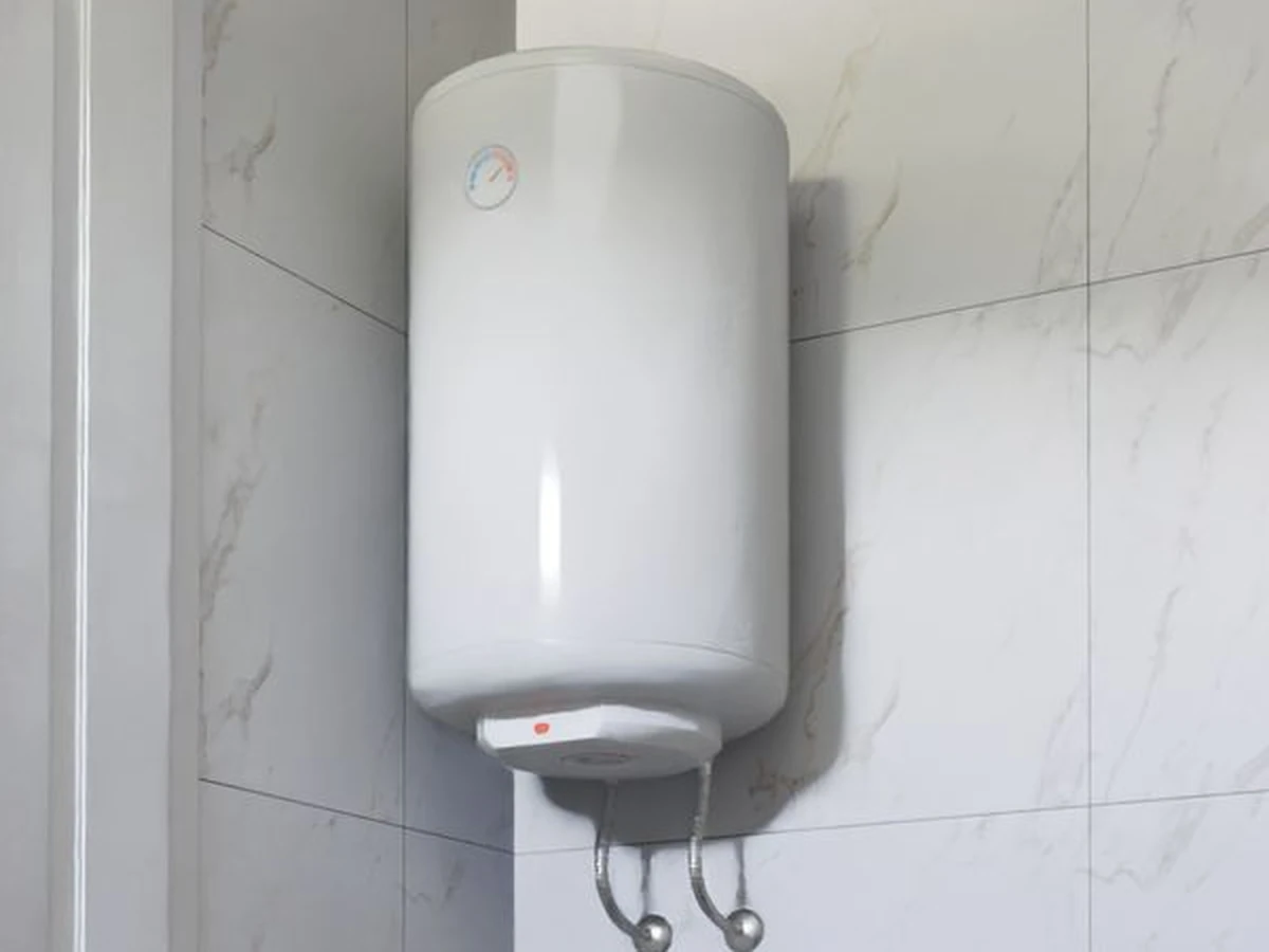 White wall-mounted domestic hot water tank with a temperature gauge and flexible pipe connections, on a pale tiled wall